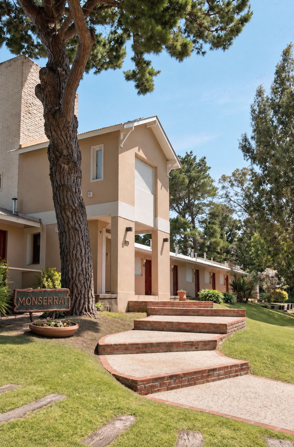 Bungalow dúplex de dos plantas junto a un pino, con escalones de ladrillo y el cartel de Monserrat
