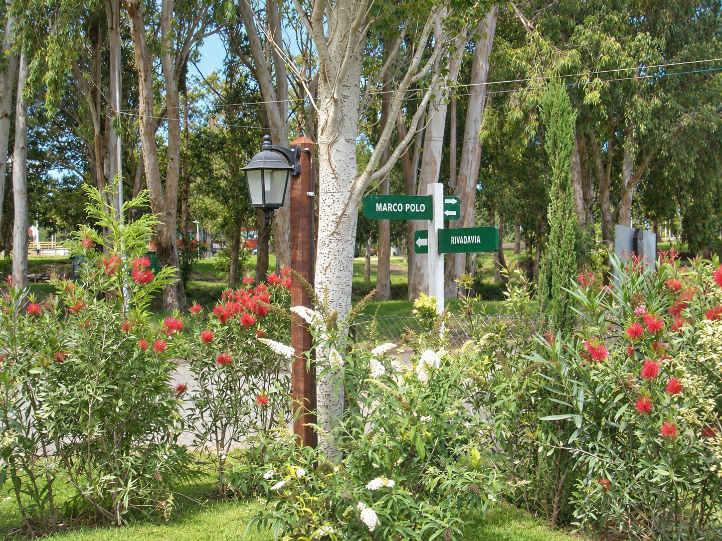 Esquina del complejo con flores rojas y blancas, un farol y el cartel de las calles Rivadavia y Marco Polo entre los árboles